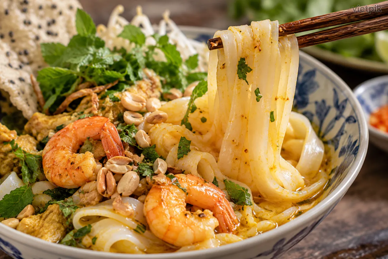 Wide rice noodles lifted above a bowl of mì Quảng